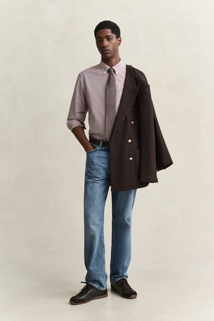 A man modeling a dark brown double-breasted blazer draped over his right shoulder, worn over a light purple button-down shirt with a patterned tie, blue jeans, and dark brown leather shoes, against a plain beige studio background.