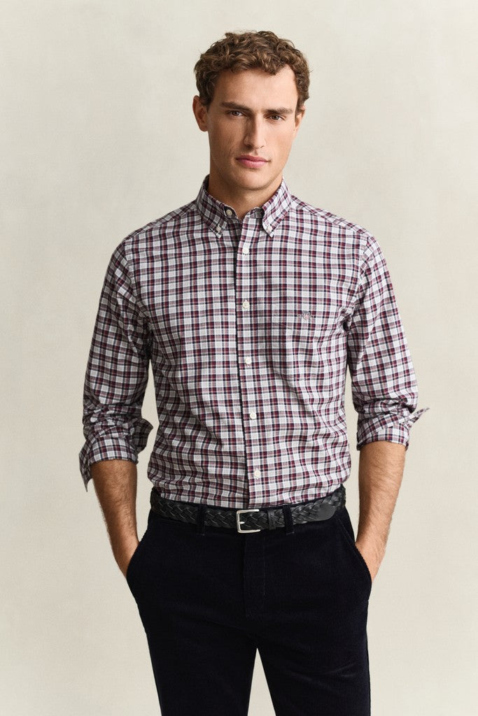 A man wearing a long-sleeve button-down shirt with a red, white, and dark blue plaid pattern, paired with black trousers and a black belt, standing against a light beige background.