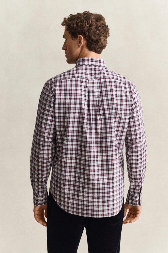 A man viewed from the back wearing a long-sleeve plaid button-up shirt with a red, white, and dark check pattern, paired with dark pants, against a plain light beige background.