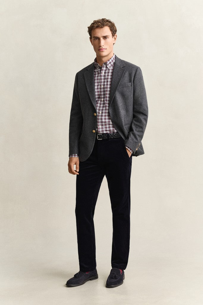 A man modeling a grey blazer over a red and white checkered shirt, paired with black trousers and dark loafers, standing against a neutral beige studio background.