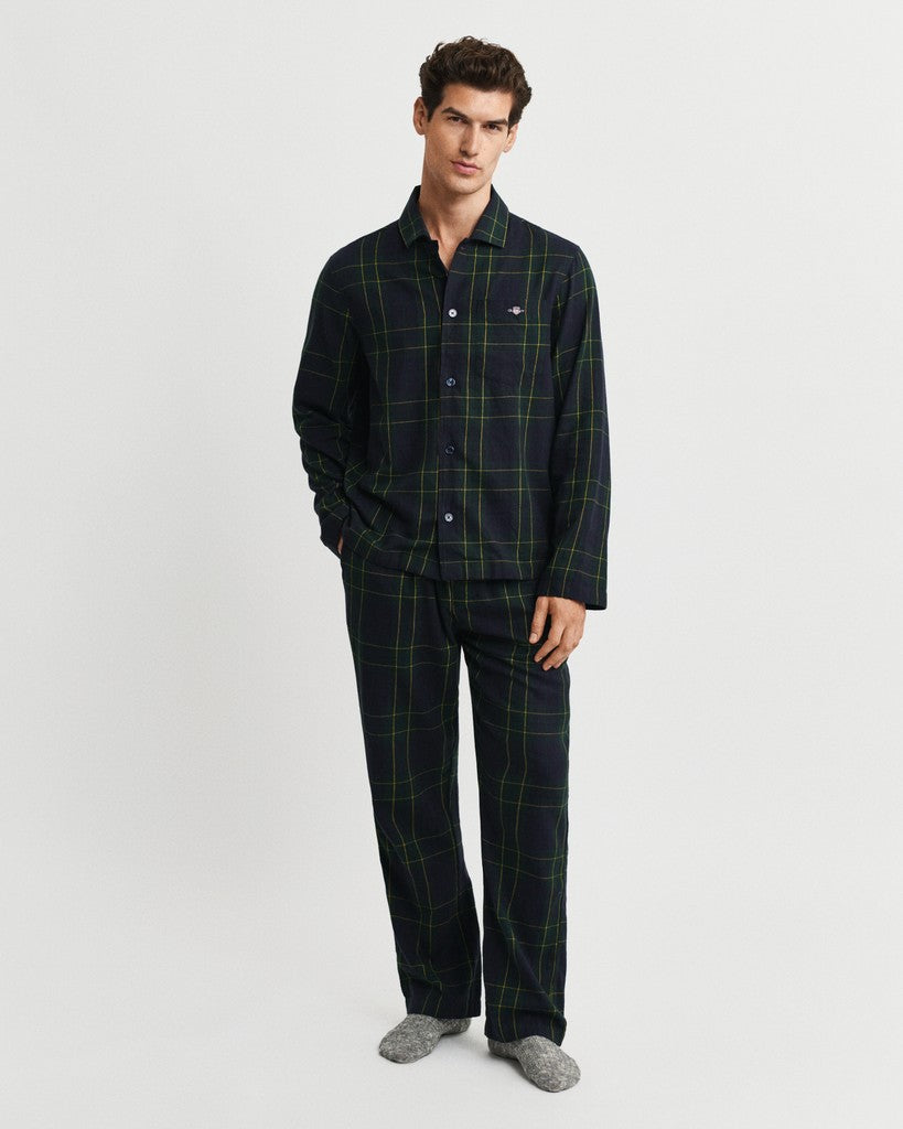 A man modeling a two-piece pajama set consisting of a long-sleeve button-up shirt and matching pants in a dark navy blue plaid pattern with thin green lines, paired with gray socks, against a light gray studio background.