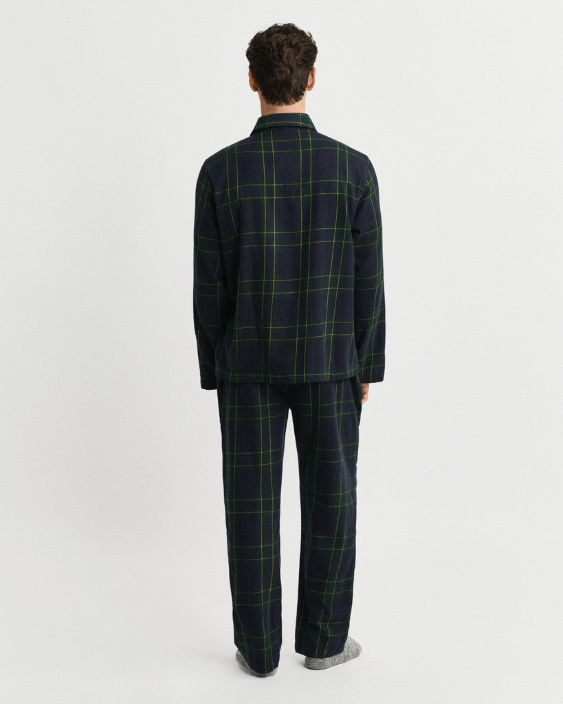 A man viewed from the back wearing a dark navy plaid pajama set with green windowpane lines, consisting of a long-sleeve top and matching trousers, against a plain light gray background.
