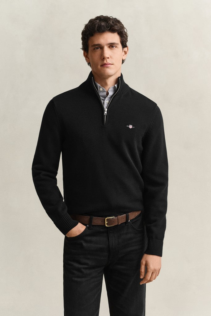A man wearing a black quarter-zip knit sweater with a small embroidered Gant logo on the left chest, layered over a light blue collared shirt and paired with dark trousers and a brown belt, standing against a neutral light gray background.