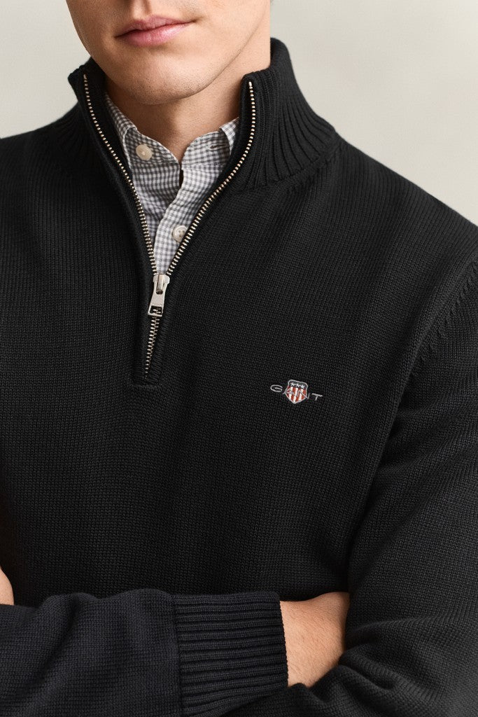 A close-up of a man wearing a black ribbed quarter-zip sweater with a small embroidered GANT shield logo on the left chest, layered over a checkered shirt.