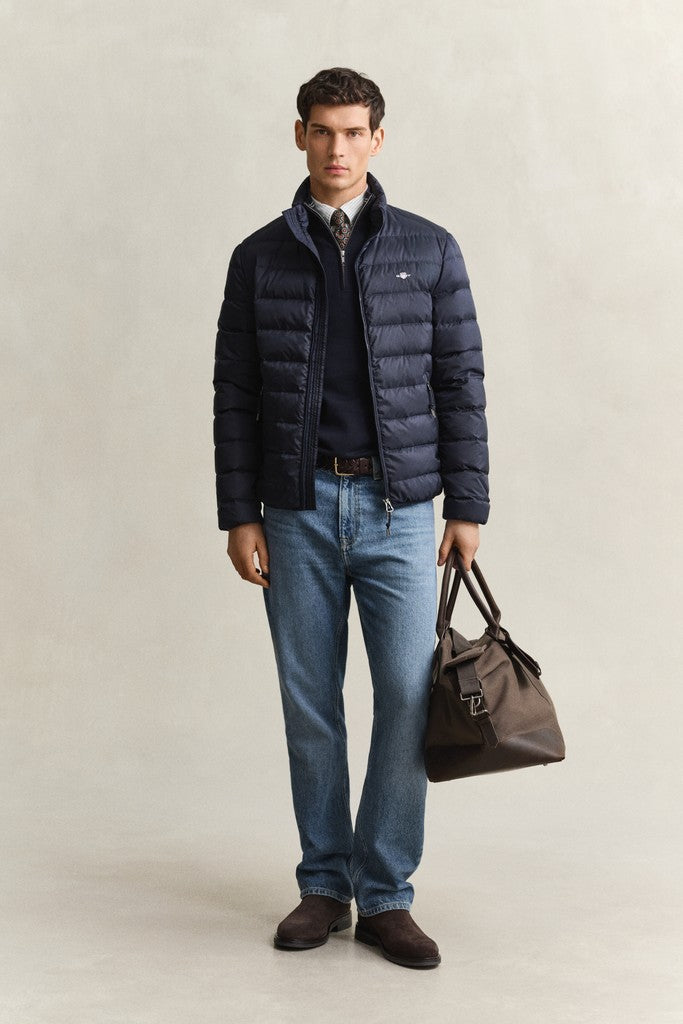 Man wearing a navy blue GANT puffer jacket with navy sweater, blue jeans, brown shoe and holding a brown travel bag