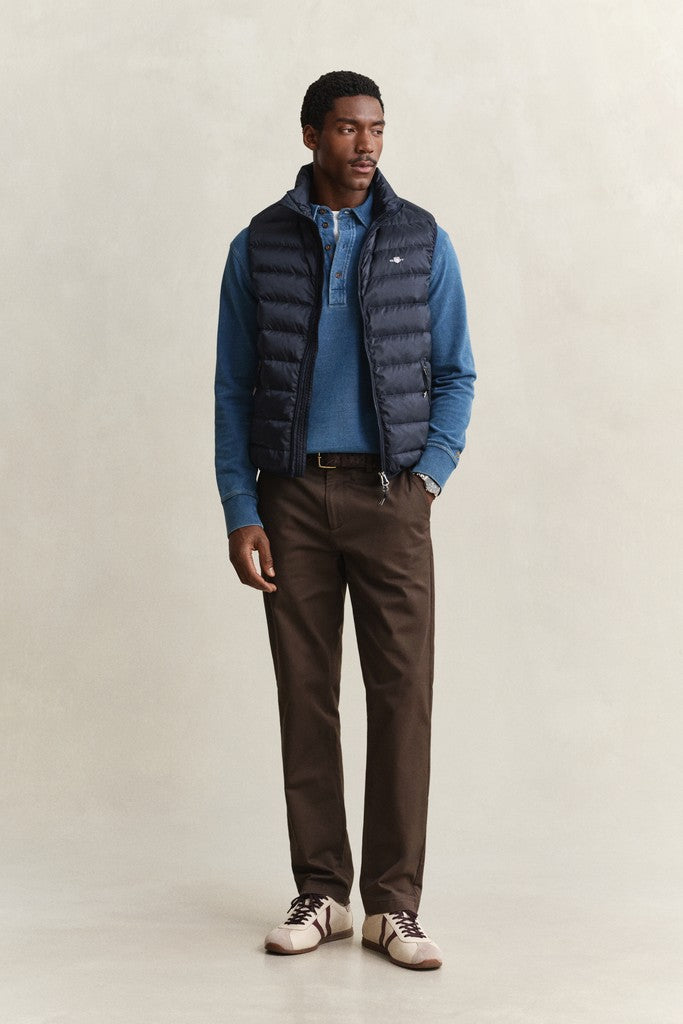 A man modeling a dark navy puffer vest over a blue polo shirt, paired with brown trousers and beige sneakers, standing against a neutral beige studio backdrop.