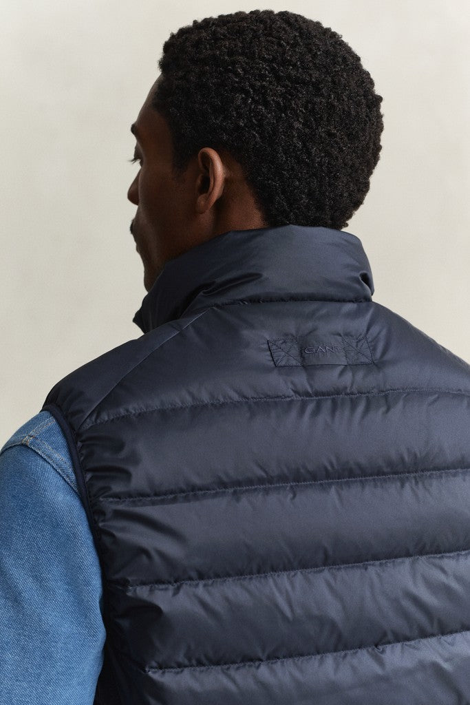 A man shown from the back wearing a navy blue quilted puffer vest over a blue denim shirt, against a plain white background.