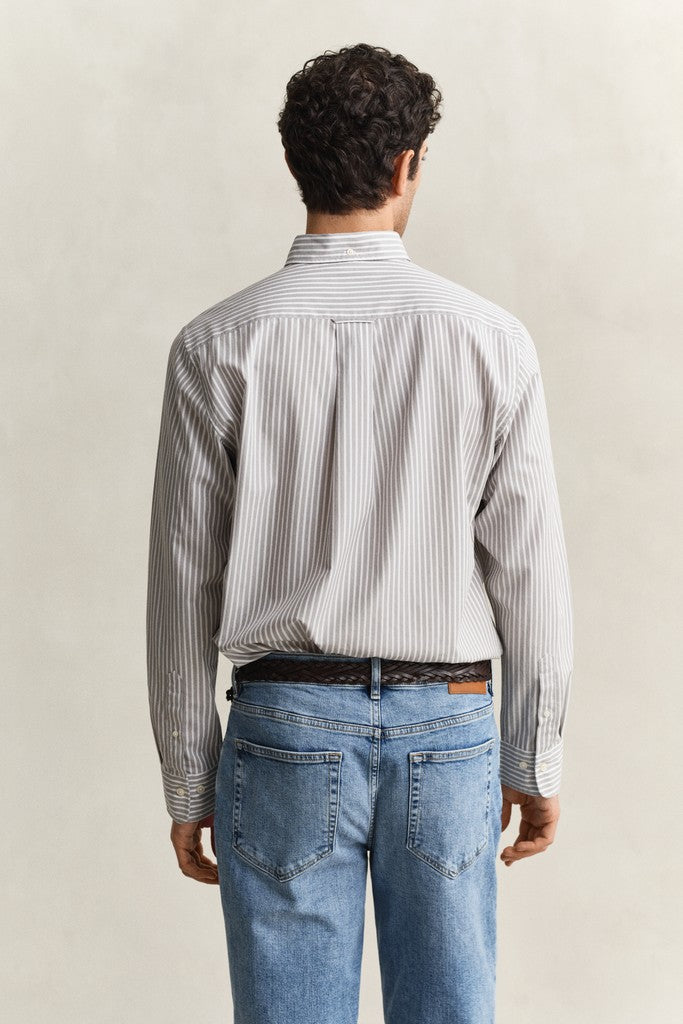 A man viewed from behind wearing a light gray and white vertically striped button-down shirt, blue jeans, and a brown belt, set against a plain beige studio background.