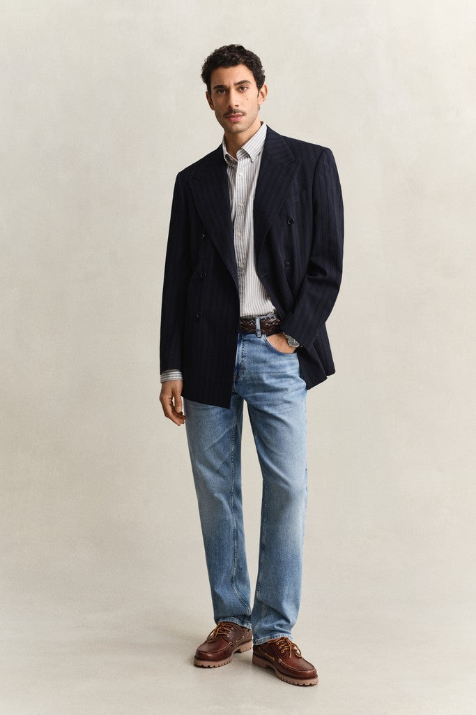Man wearing a dark blazer, light shirt, blue jeans, and brown shoes on a beige background