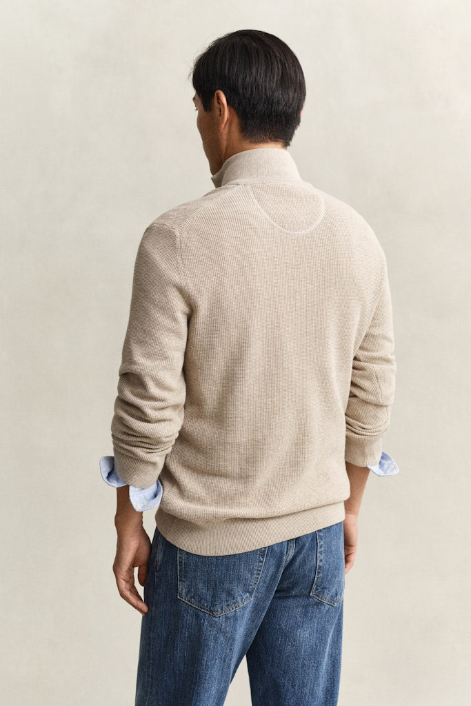 Man wearing a beige sweater and blue jeans against a plain background