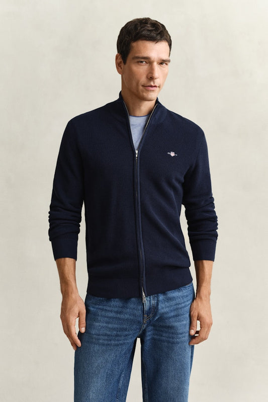 Man wearing a navy zipped cardigan with Gant logo and blue jeans against a grey background