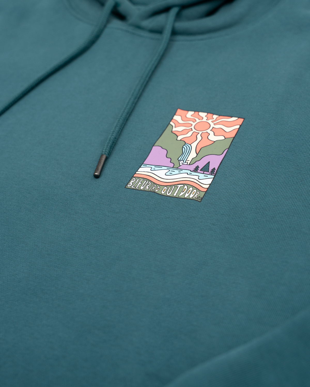 A close-up of a teal hoodie featuring a colorful rectangular chest graphic with a stylized sun, mountains, and the text 'Bleubird Outdoors'.