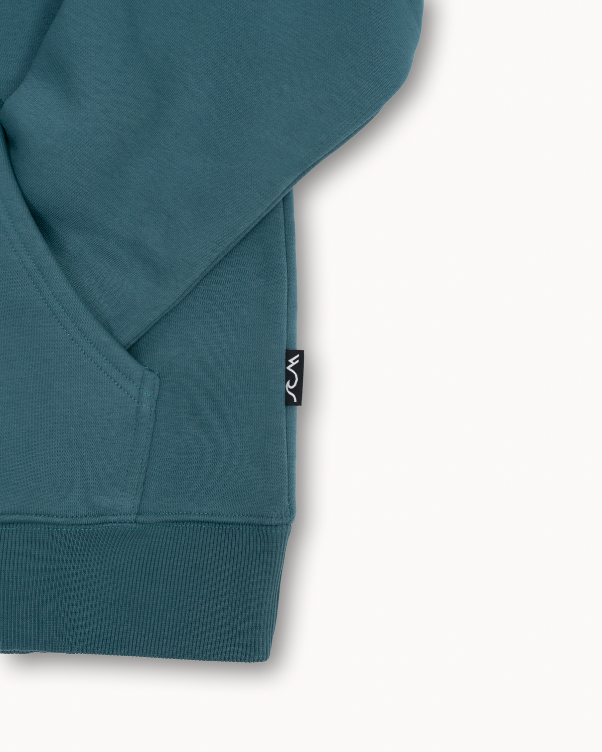 Close-up of a teal hoodie sleeve with a ribbed cuff and a small black brand tag on the side, set against a clean off-white background.