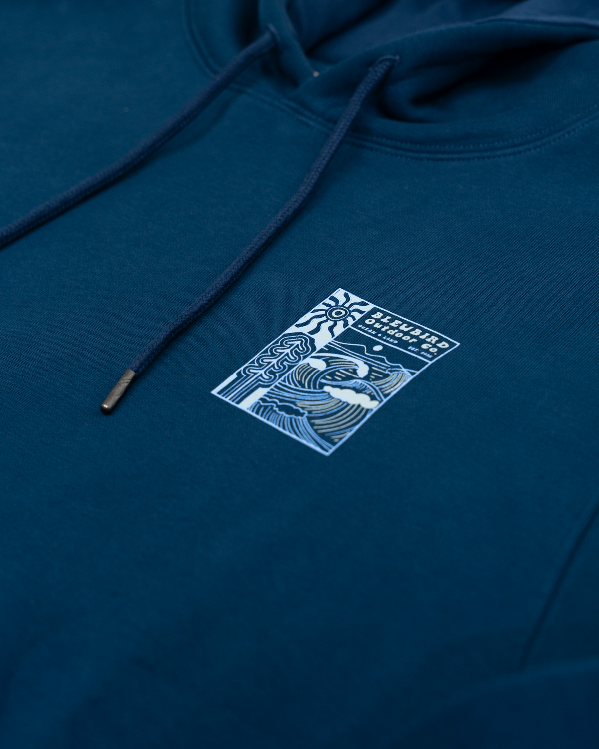 A close-up of a dark teal blue hoodie featuring a white graphic print on the chest with the text 'BLEUBIRD OUTDOOR CO' and stylized wave illustrations. The hoodie has matching dark teal drawstrings.