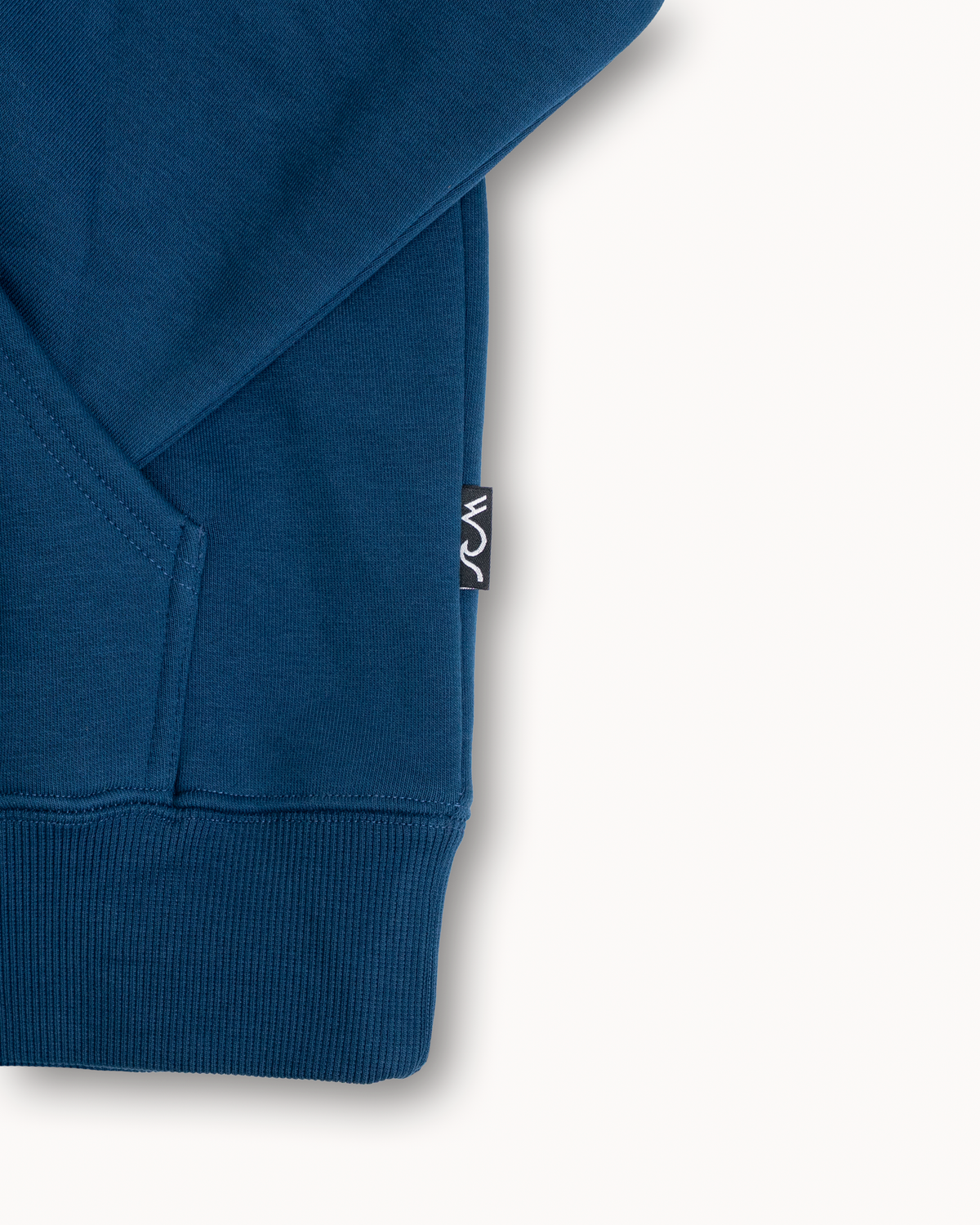 Close-up of a blue ribbed knit garment corner with a small black-and-white woven brand tag on the sleeve, photographed on a clean off-white background.
