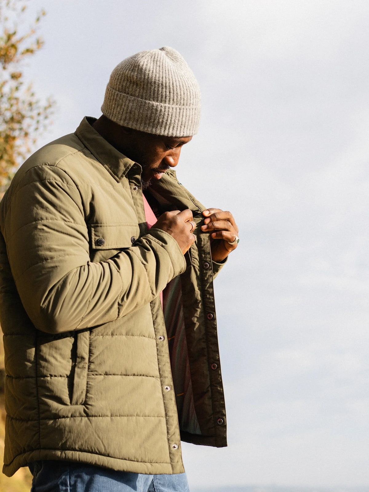 A man wearing a light beige knit beanie and an olive green quilted jacket, looking down as he adjusts the collar of the jacket outdoors against a bright sky.