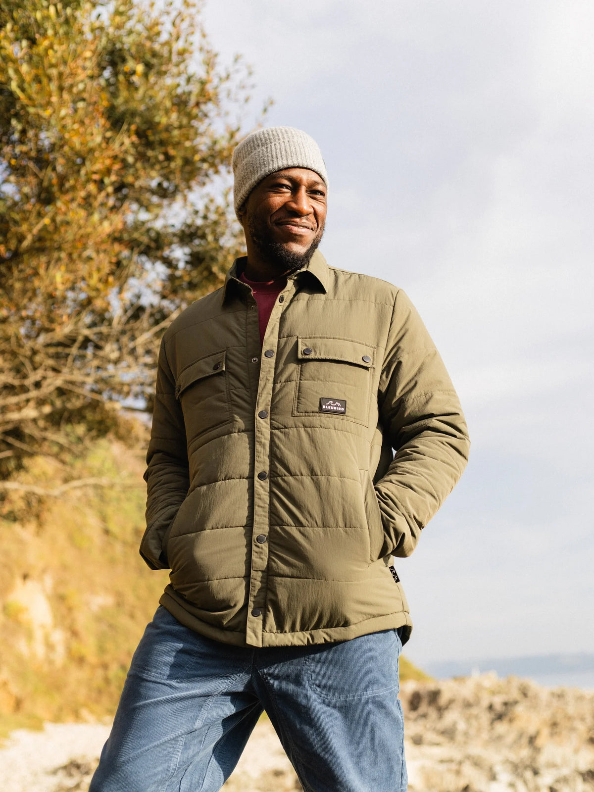 A man wearing an olive green quilted jacket, blue jeans, and a white knit beanie stands outdoors on a rocky path with trees in the background.