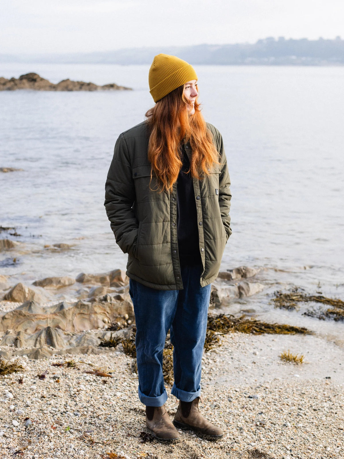 A woman with long red hair stands on a rocky shoreline wearing an olive green quilted jacket, blue jeans, brown boots, and a mustard yellow beanie, looking off-camera toward the water.