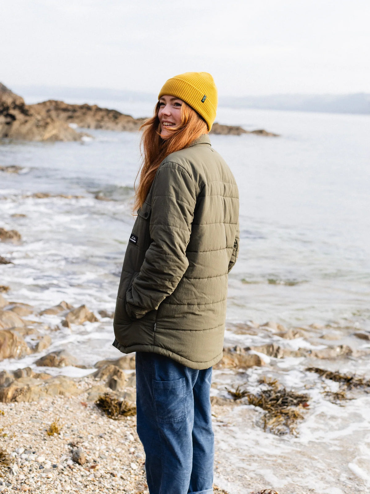 A woman standing on a rocky beach wearing an olive green quilted puffer jacket, blue jeans, and a bright yellow knit beanie, looking back over her shoulder.