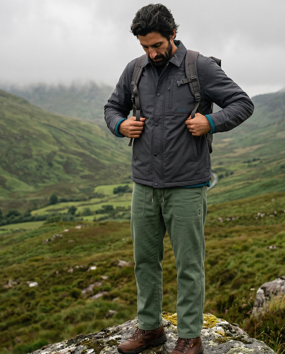 A man standing on a rocky mountain path wearing a dark gray button-up jacket, olive green pants, and brown hiking boots, with a backpack on, against a backdrop of green hills under a cloudy sky.