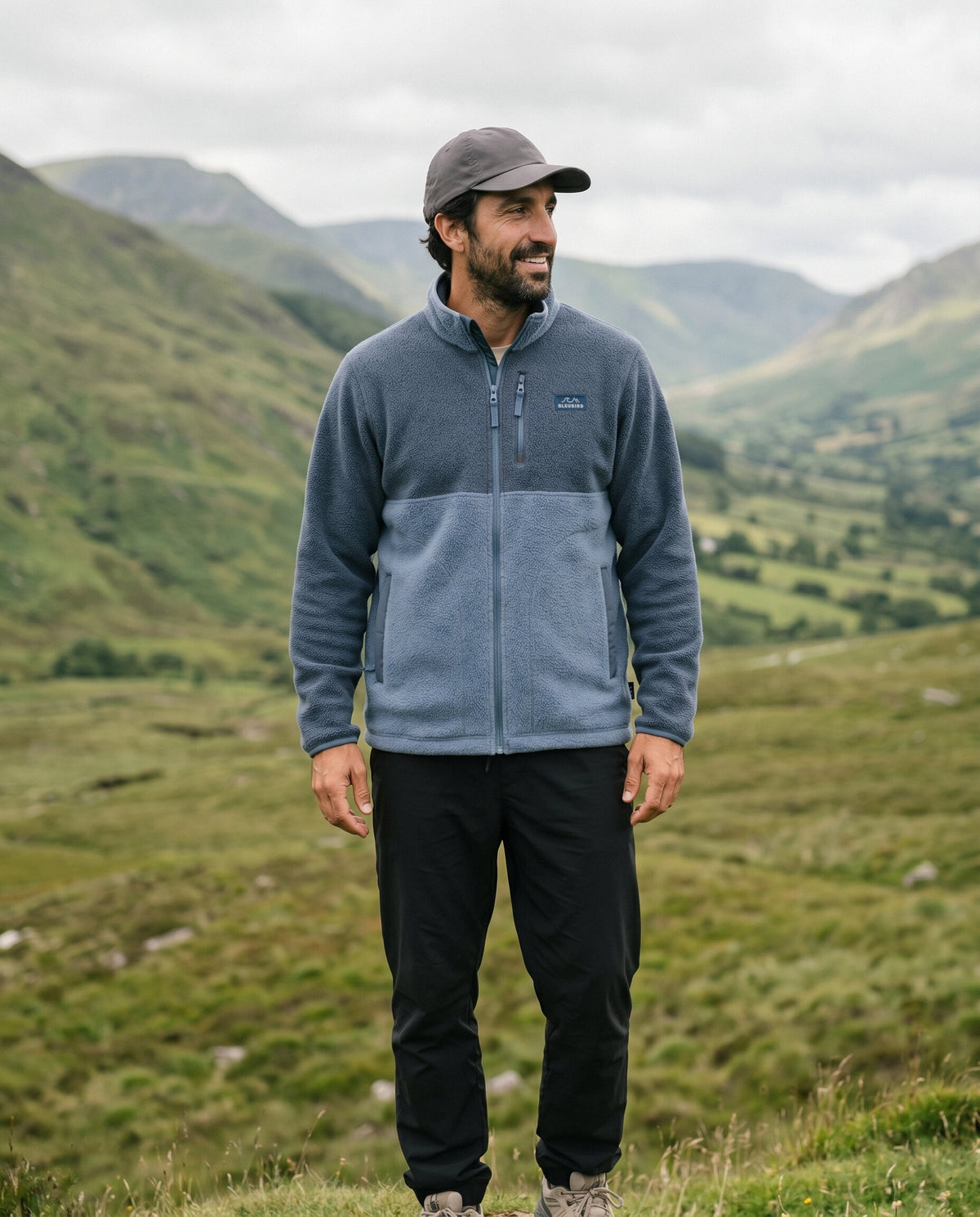 A man wearing a Bleubird fleece pullover in slate blue and heather blue, black pants, and a dark cap, standing in a grassy mountain valley under an overcast sky.