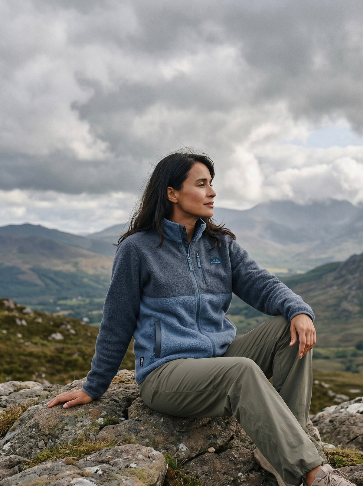 A woman sits on a rocky mountain peak wearing a slate blue and gray fleece zip-up jacket and olive green pants, with a dramatic cloudy sky and distant mountains in the background.