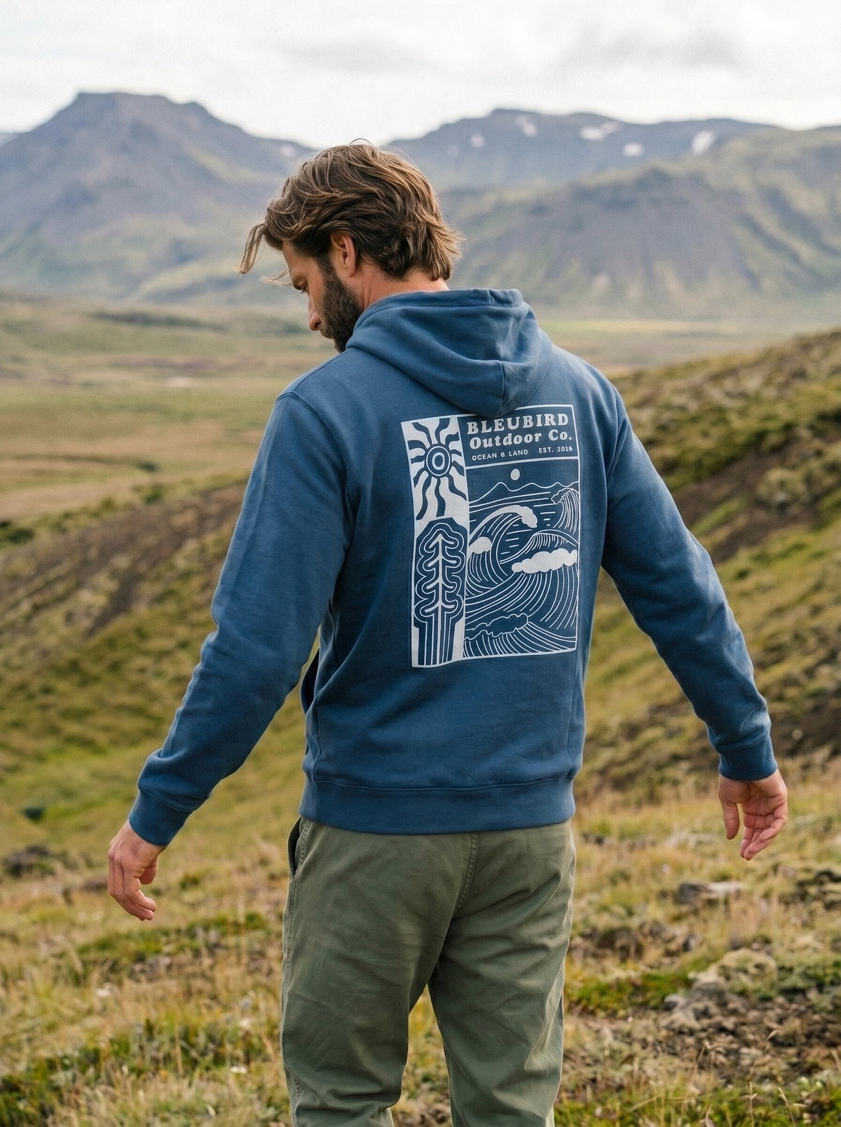 A man standing outdoors in a grassy mountain landscape, viewed from behind, wearing a blue hoodie with a large white graphic print on the back that reads 'BLEUBIRD Outdoor Co.' and olive green pants.