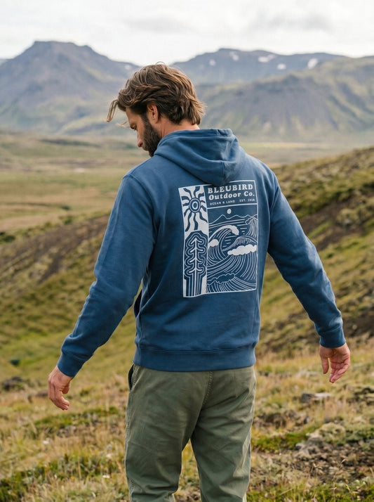 A man standing outdoors in a grassy mountain landscape, viewed from behind, wearing a blue hoodie with a large white graphic print on the back that reads 'BLEUBIRD Outdoor Co.' and olive green pants.