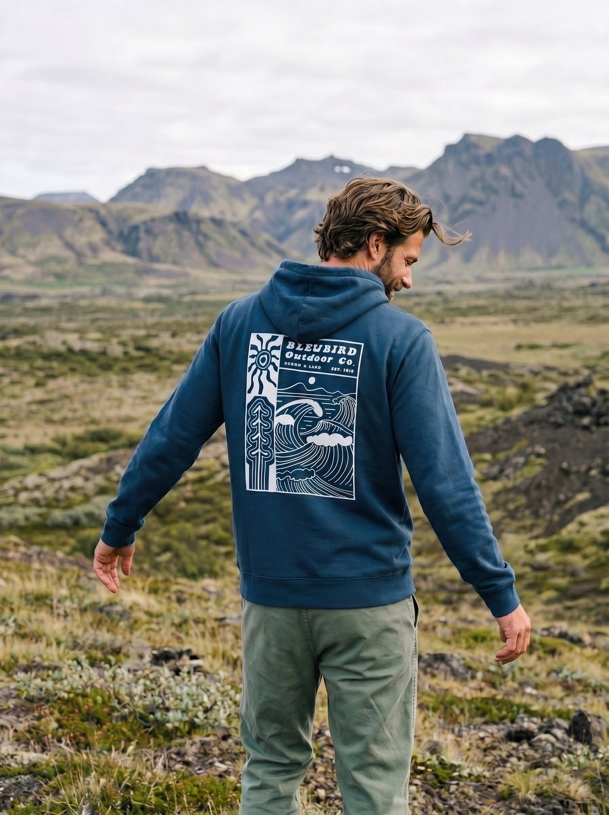 A man standing outdoors in a mountain landscape, viewed from behind, wearing a navy blue hoodie with a large white graphic print on the back that reads 'BLEUBIRD OUTDOOR Co.' and olive green pants.