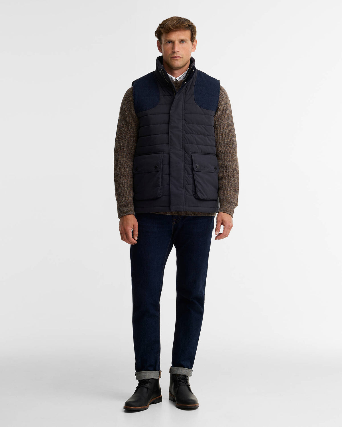 A man modeling a dark navy quilted puffer vest over a brown knit sweater, paired with dark blue jeans and black boots, against a plain white studio background.