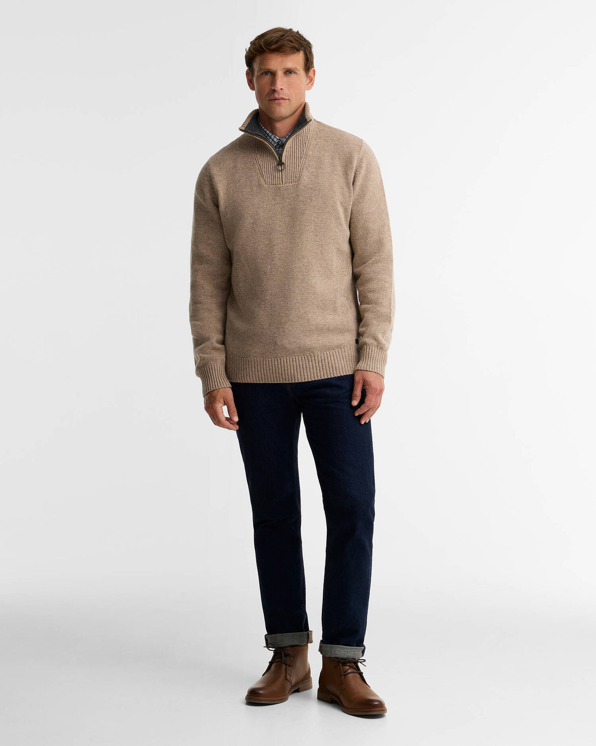 A man modeling a light beige quarter-zip knit sweater with a dark collar, paired with navy blue jeans and brown leather boots, against a plain white studio background.