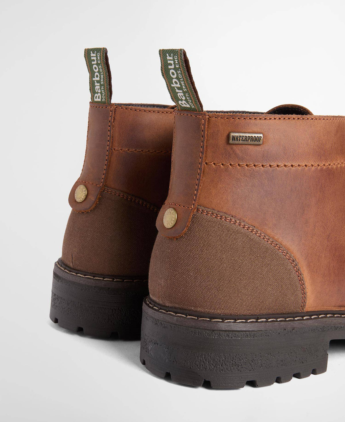 Close-up rear view of a pair of brown leather Barbour boots with dark rubber soles and green pull tabs on a light gray background.
