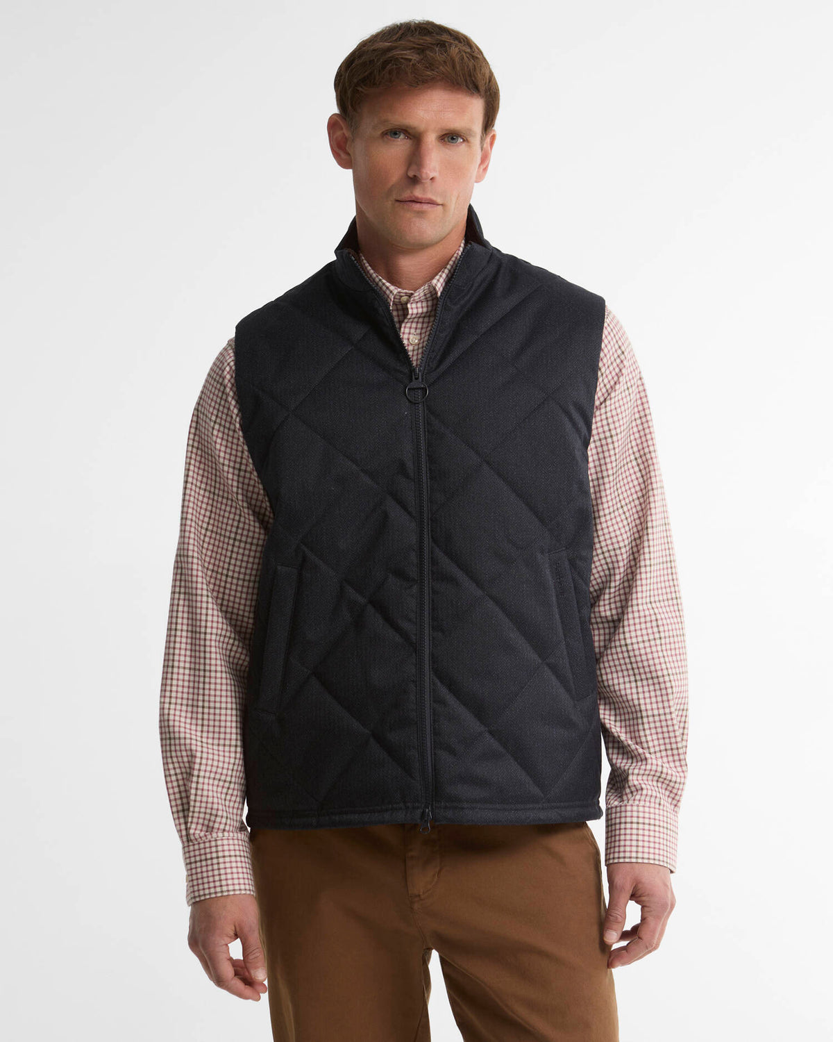 A man wearing a navy diamond-quilted zip-up vest over a pink and white checkered button-down shirt, paired with brown pants, photographed against a light gray studio background.