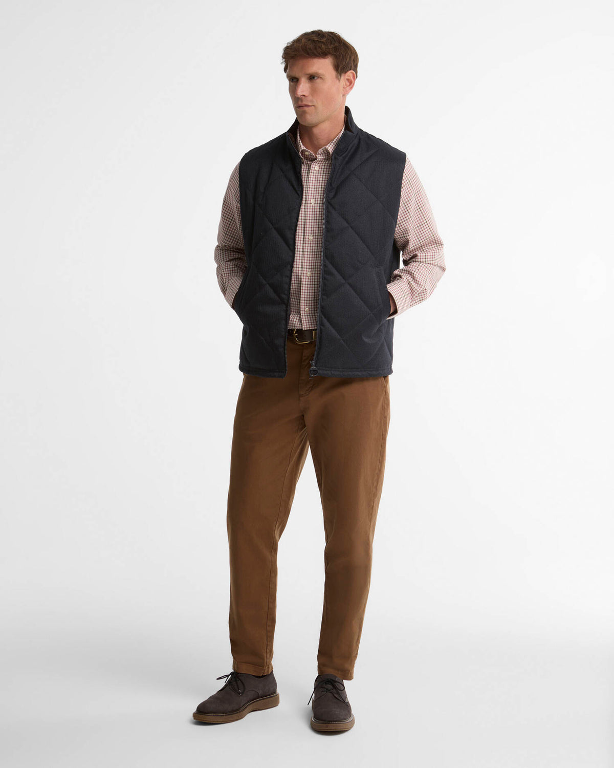 A man modeling a dark navy quilted vest over a pinkish-beige checkered shirt, paired with brown trousers and dark brown shoes, standing against a plain white studio background.