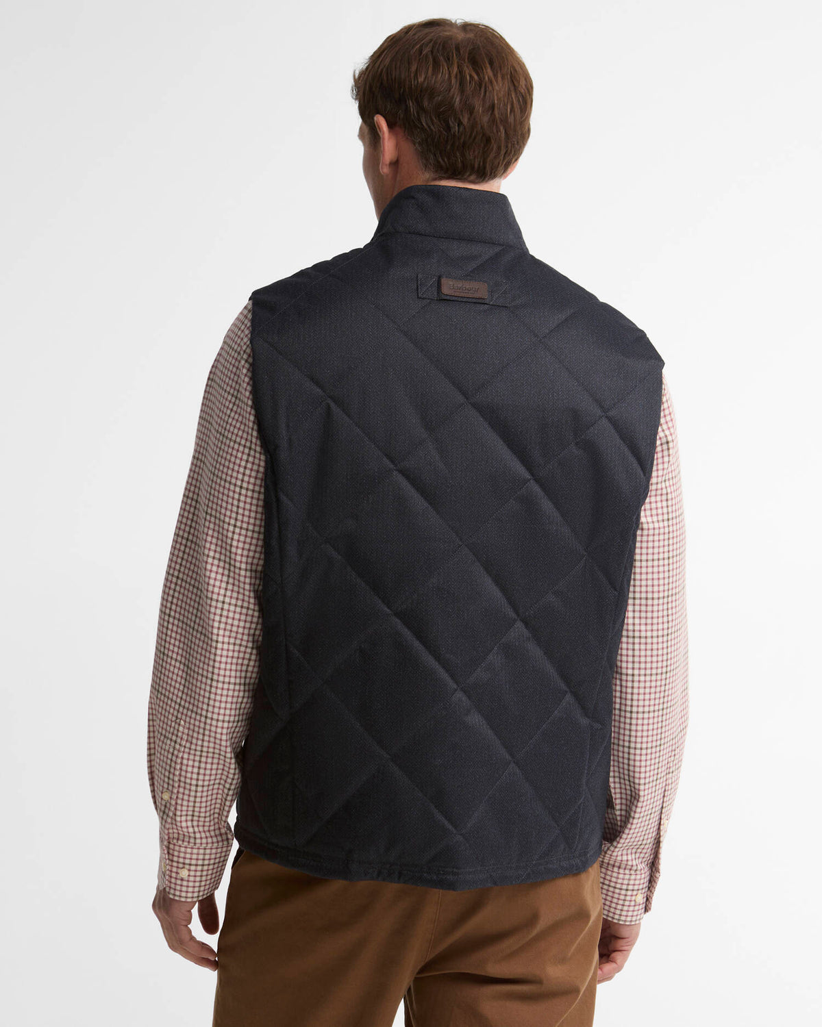 A man viewed from the back wearing a dark navy quilted vest over a beige and white checkered shirt, paired with brown trousers, against a plain white studio background.