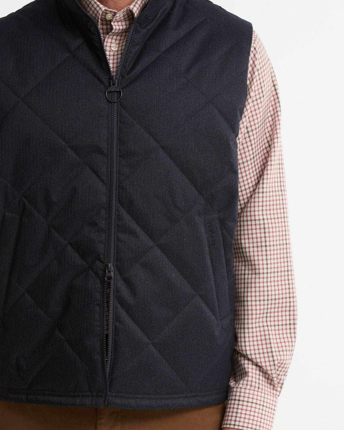 A close-up of a person wearing a navy diamond-quilted zip-front vest over a red and white gingham check shirt, with brown pants visible at the bottom, against a plain light background.