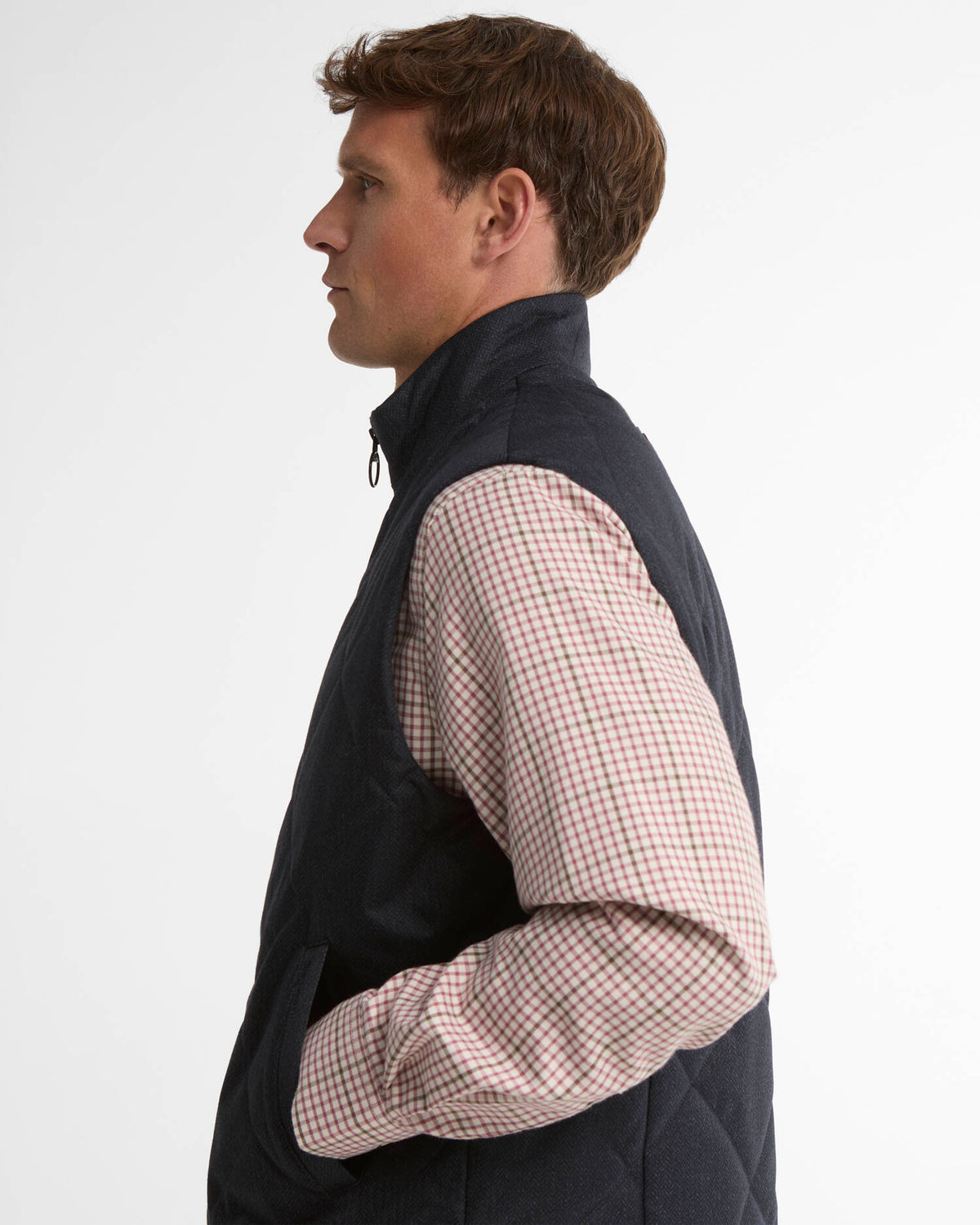 A man in profile wearing a dark navy quilted vest over a light pink and green gingham long-sleeve shirt, photographed against a clean white studio background.
