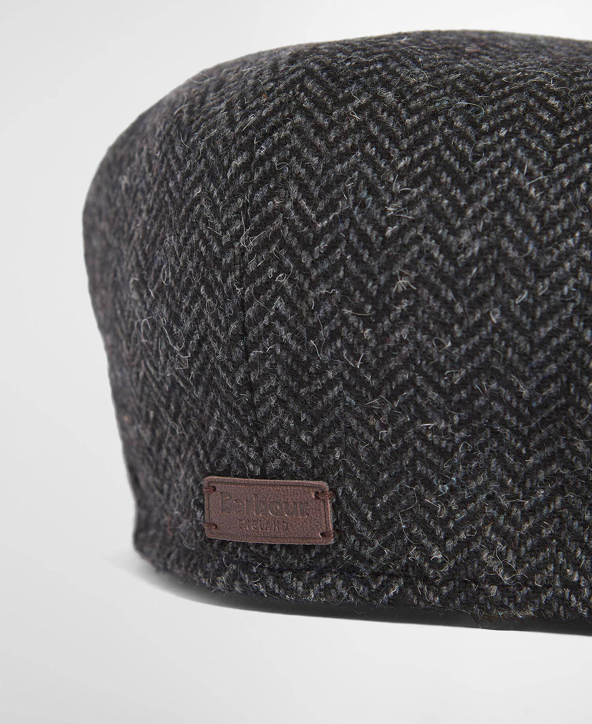 Dark gray herringbone patterned flat cap from the back with a brown leather Barbour label on a light gray background