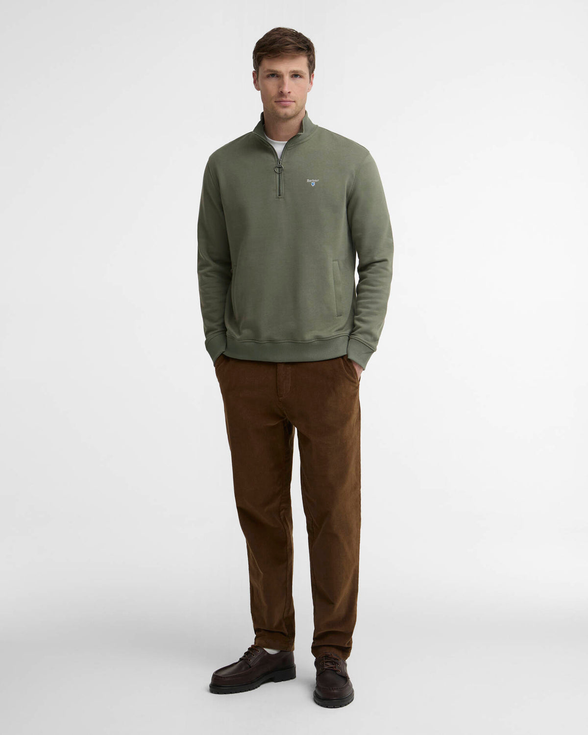 A man wearing an olive green quarter-zip pullover sweater, brown corduroy trousers, and dark brown leather shoes, standing against a light gray studio background.