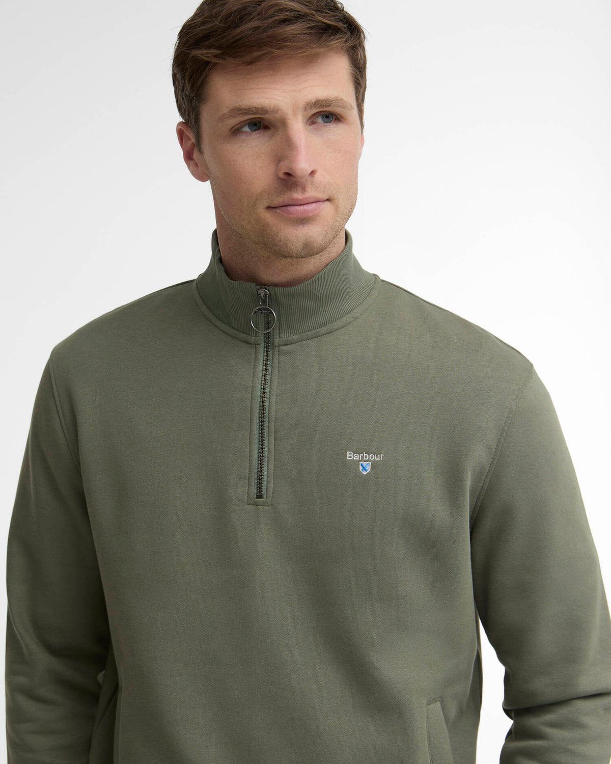 A man wearing a sage green quarter-zip pullover sweatshirt with a small Barbour logo on the left chest, photographed against a clean white studio background.