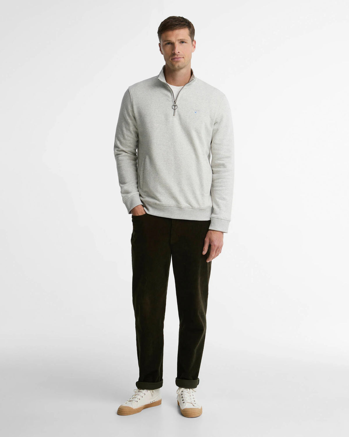 A man modeling a light gray heathered quarter-zip sweatshirt with a small embroidered logo on the chest, paired with dark olive pants and white sneakers, against a plain white studio background.