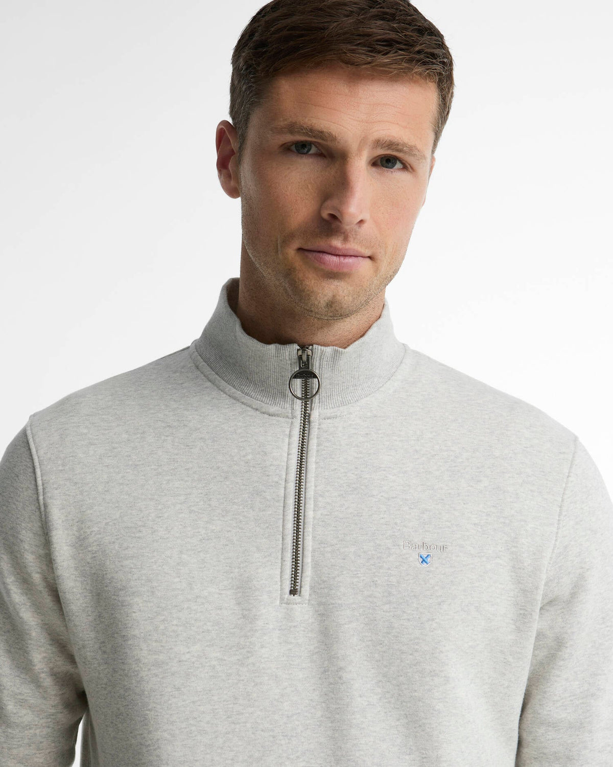 A man wearing a light gray heathered quarter-zip pullover sweatshirt with a small blue embroidered Barbour logo on the chest, photographed against a clean white studio background.