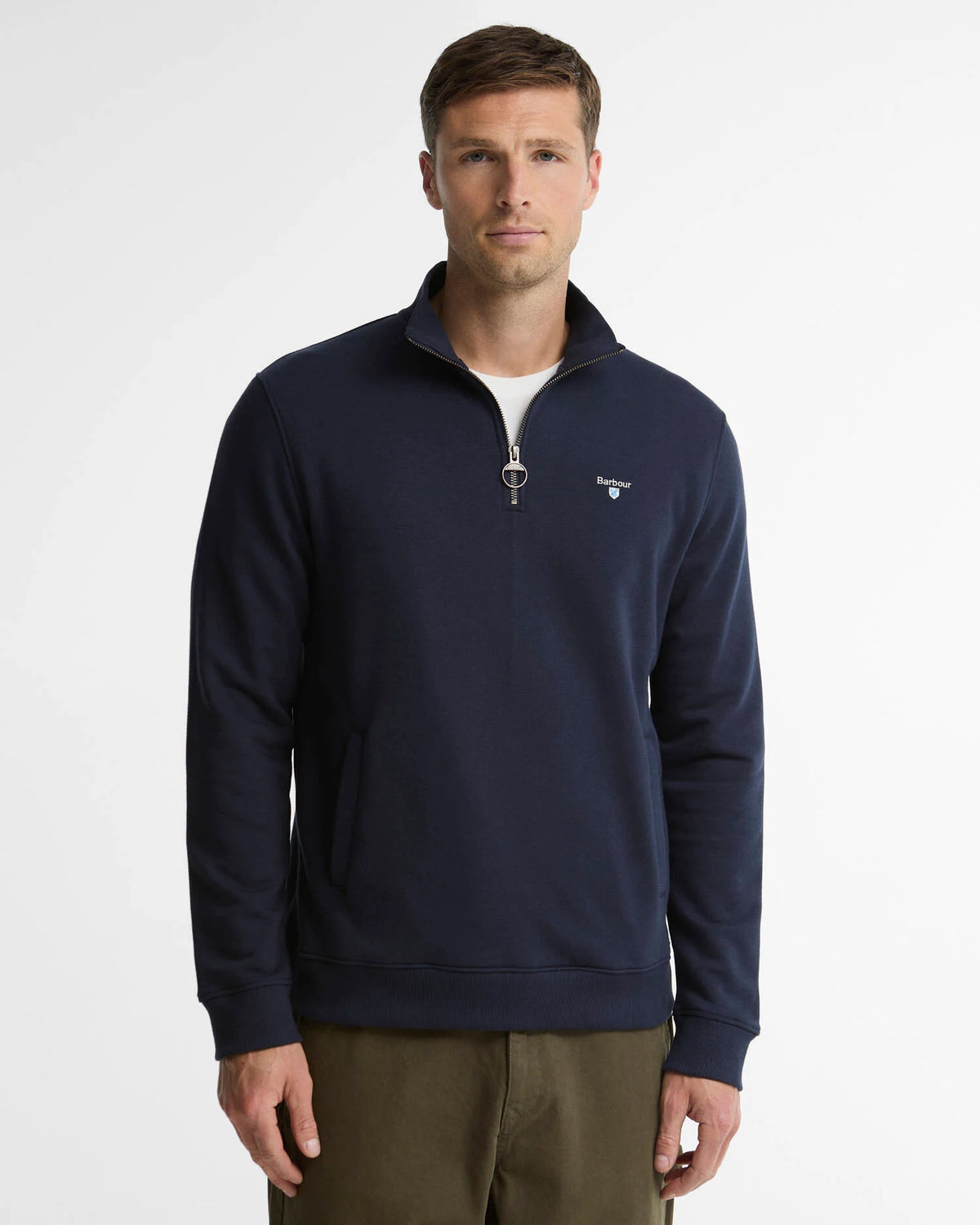 A man wearing a navy blue quarter-zip sweatshirt with a small embroidered Barbour logo on the left chest, paired with olive green pants, photographed against a light gray studio background.