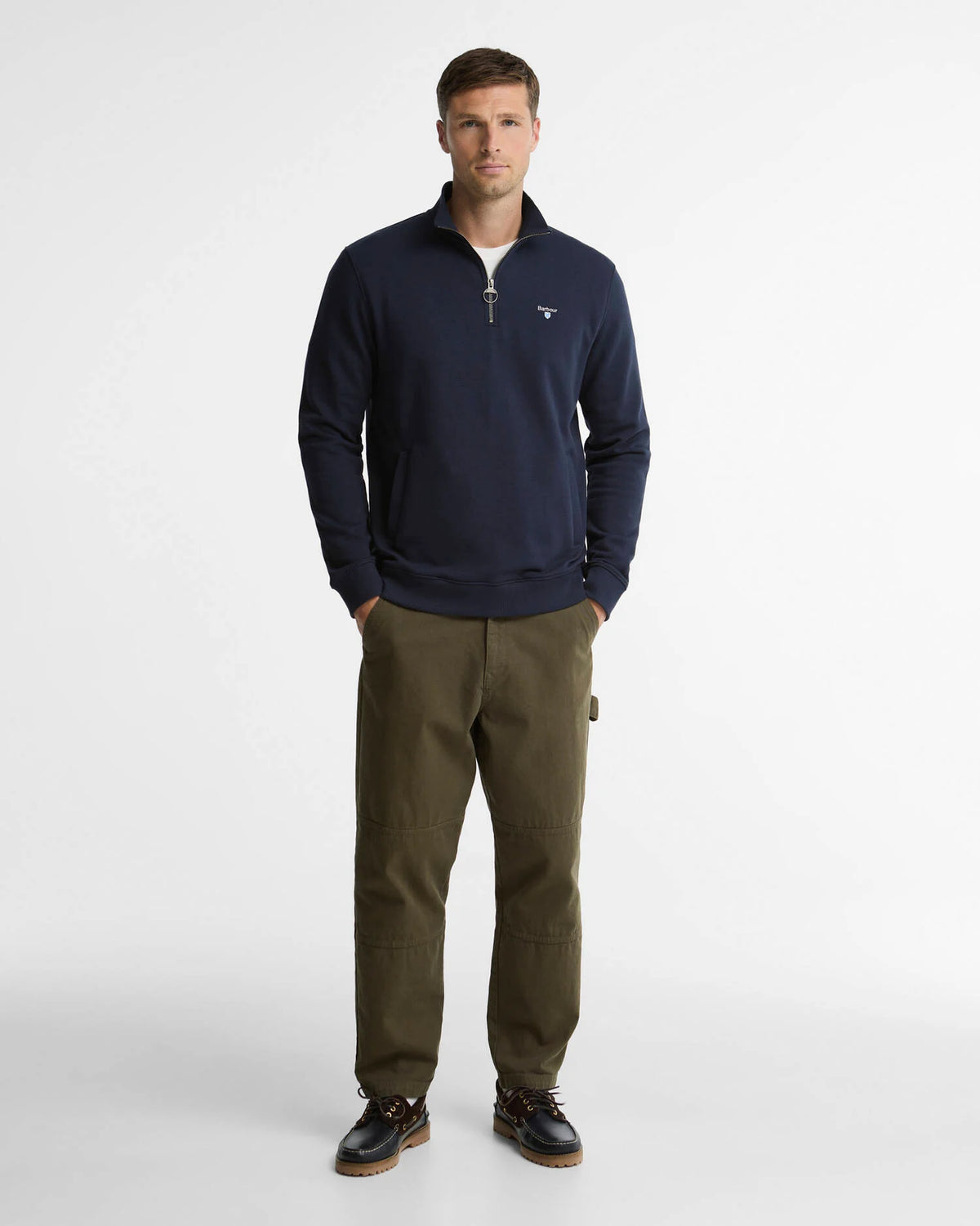A man modeling a navy blue quarter-zip pullover sweater with a small white embroidered Barbour logo on the left chest, paired with olive green trousers and dark brown shoes, against a plain light gray studio background.