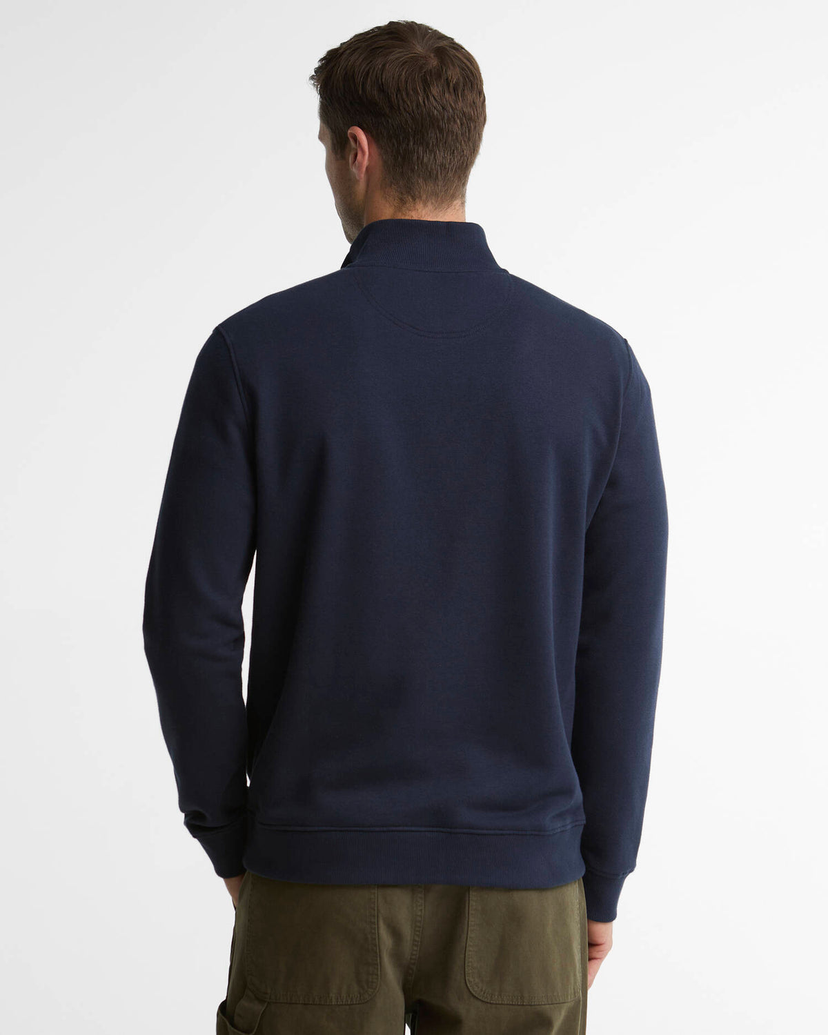 A man viewed from behind wearing a navy blue quarter-zip sweatshirt and olive green cargo pants, set against a plain white studio background.