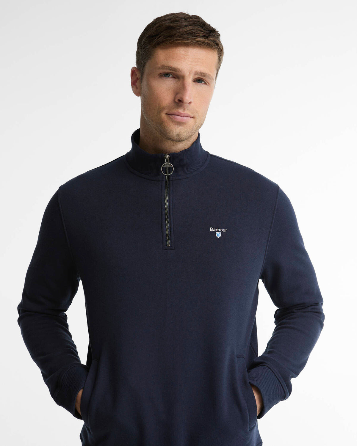 A man wearing a navy blue quarter-zip pullover sweater with a small white Barbour logo on the left chest, photographed against a light gray studio background.