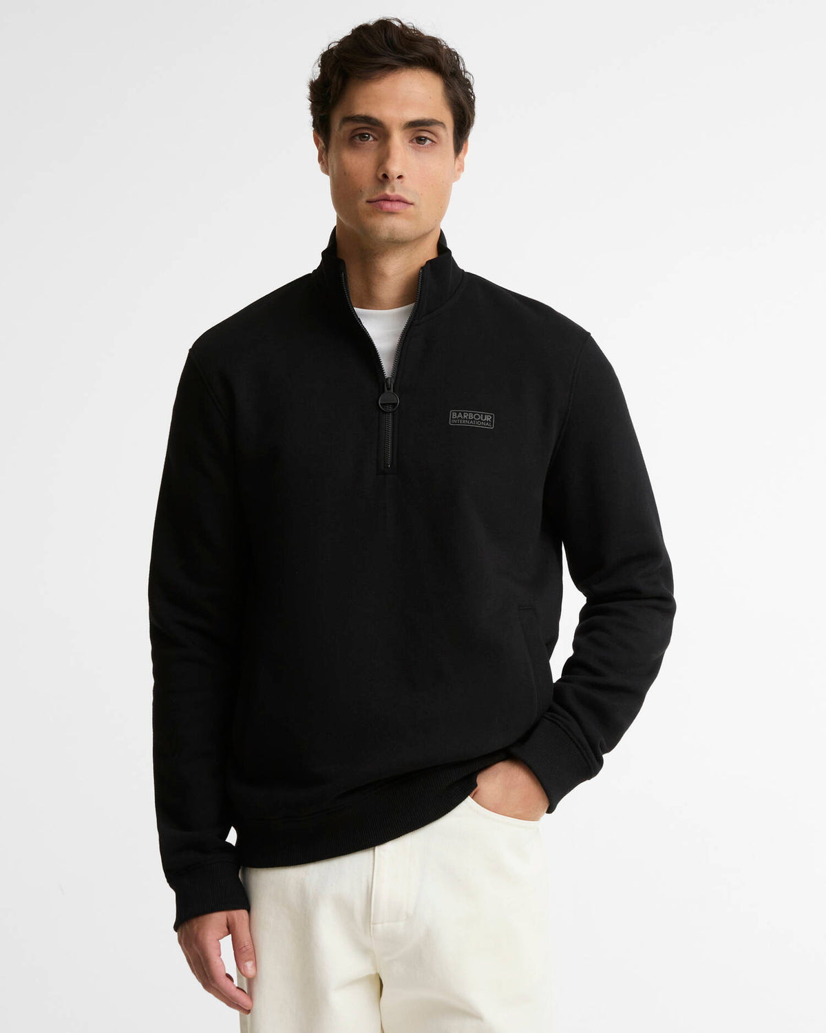 A man wearing a black quarter-zip pullover sweatshirt with a small rectangular logo patch on the left chest, paired with off-white trousers, photographed against a clean white studio background.