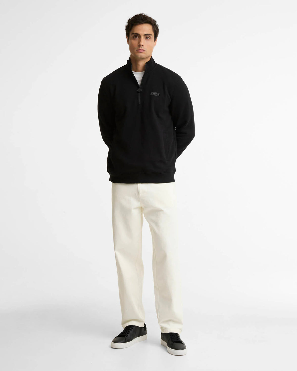 A man stands against a plain light gray studio background wearing a black quarter-zip pullover sweatshirt, off-white trousers, and black sneakers.