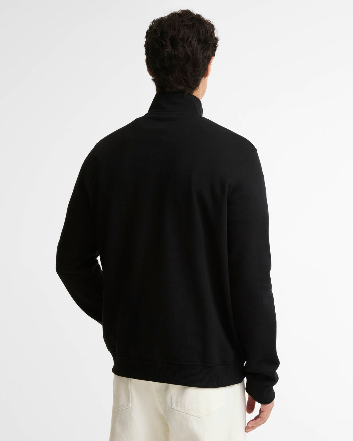 A man viewed from behind wearing a black mock neck pullover sweatshirt and off-white trousers, set against a plain light gray studio background.
