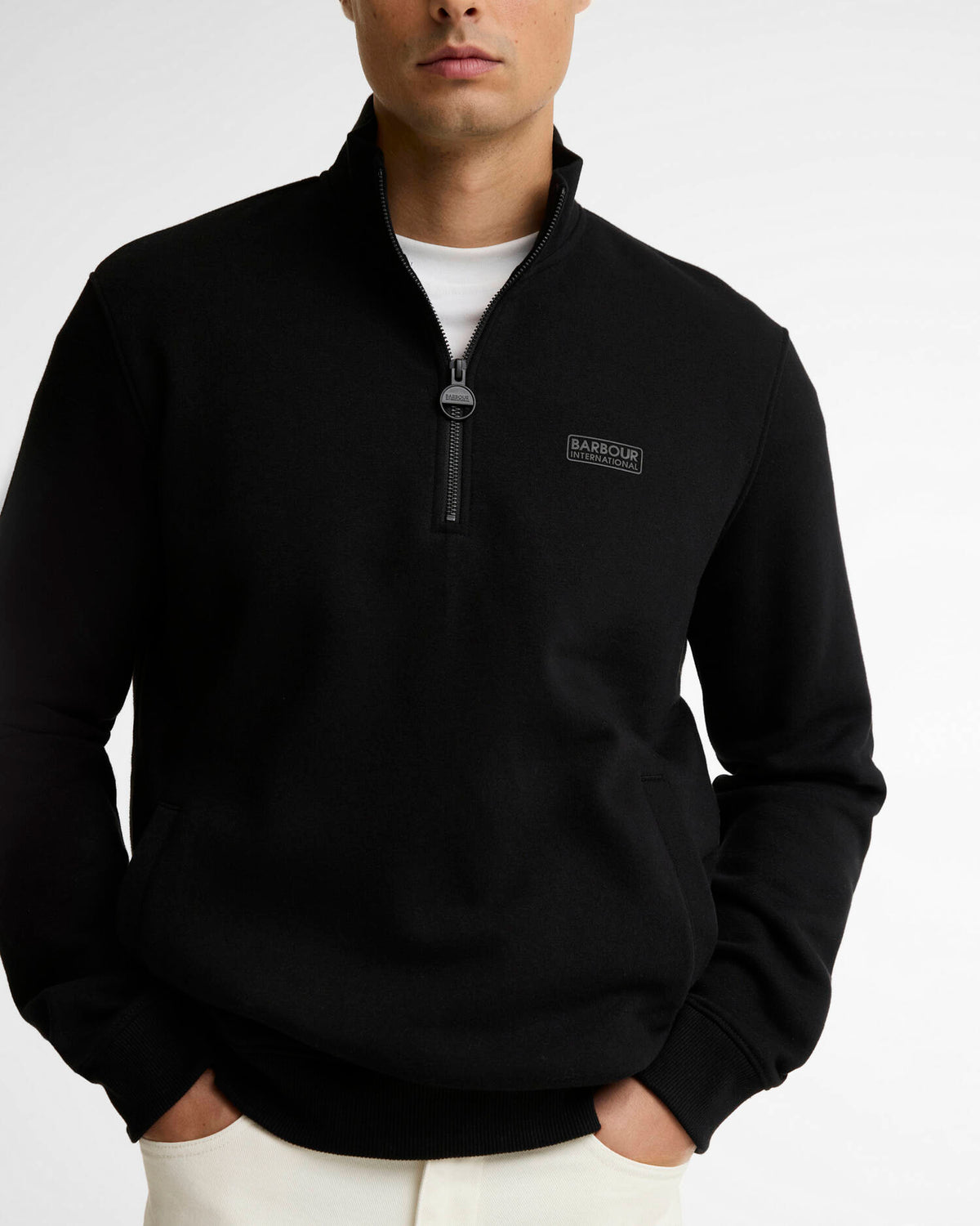 A man wearing a black quarter-zip pullover sweatshirt with a small black rectangular logo on the left chest, layered over a white t-shirt and paired with white pants, shot against a light gray studio background.